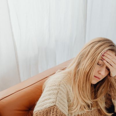 Young woman sitting on a couch holding her forehead with one hand, appearing unwell after a delayed allergic reaction